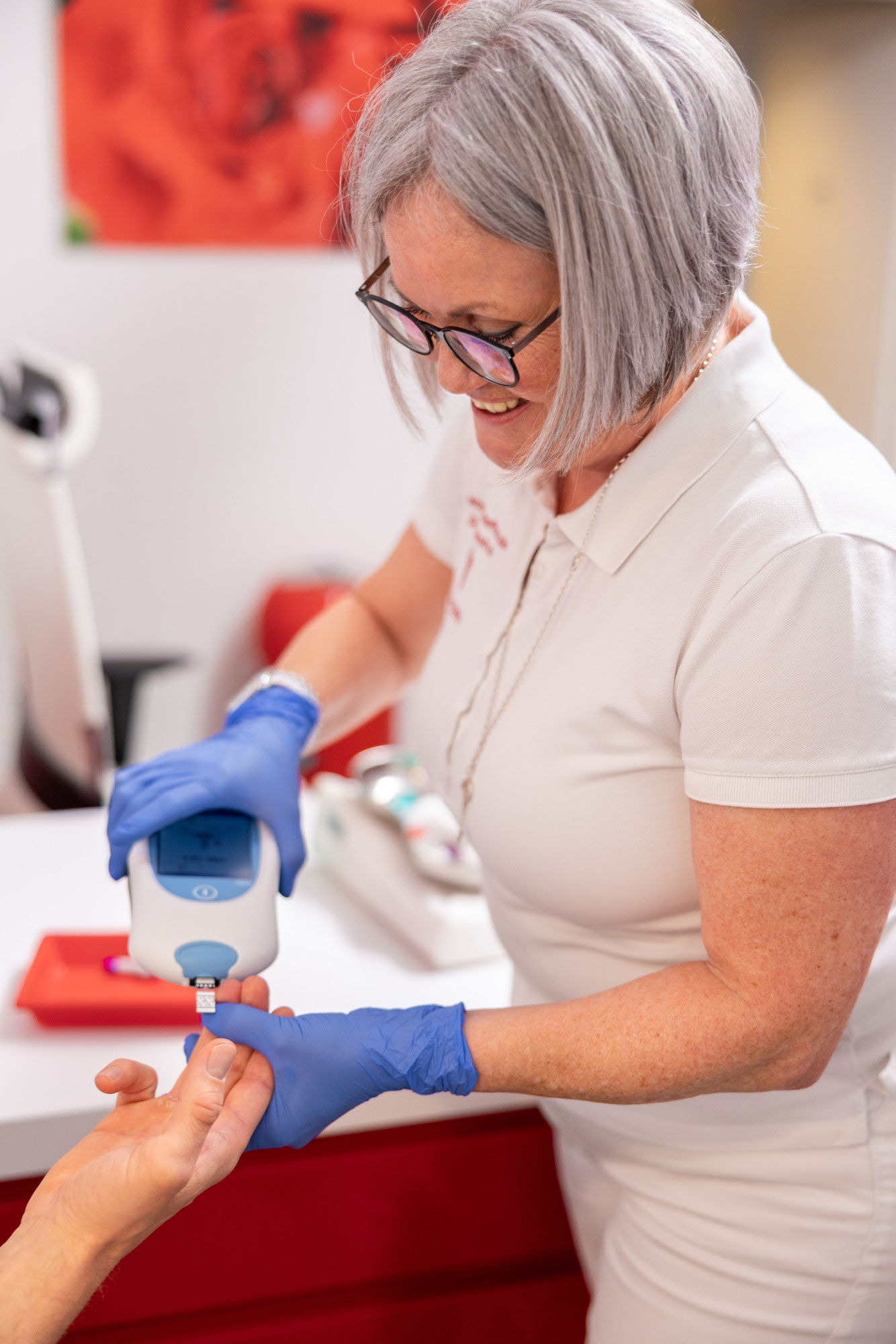 Eine lächelnde ältere Frau mit grauem Bob und Brille misst Blut mit Handschuhen