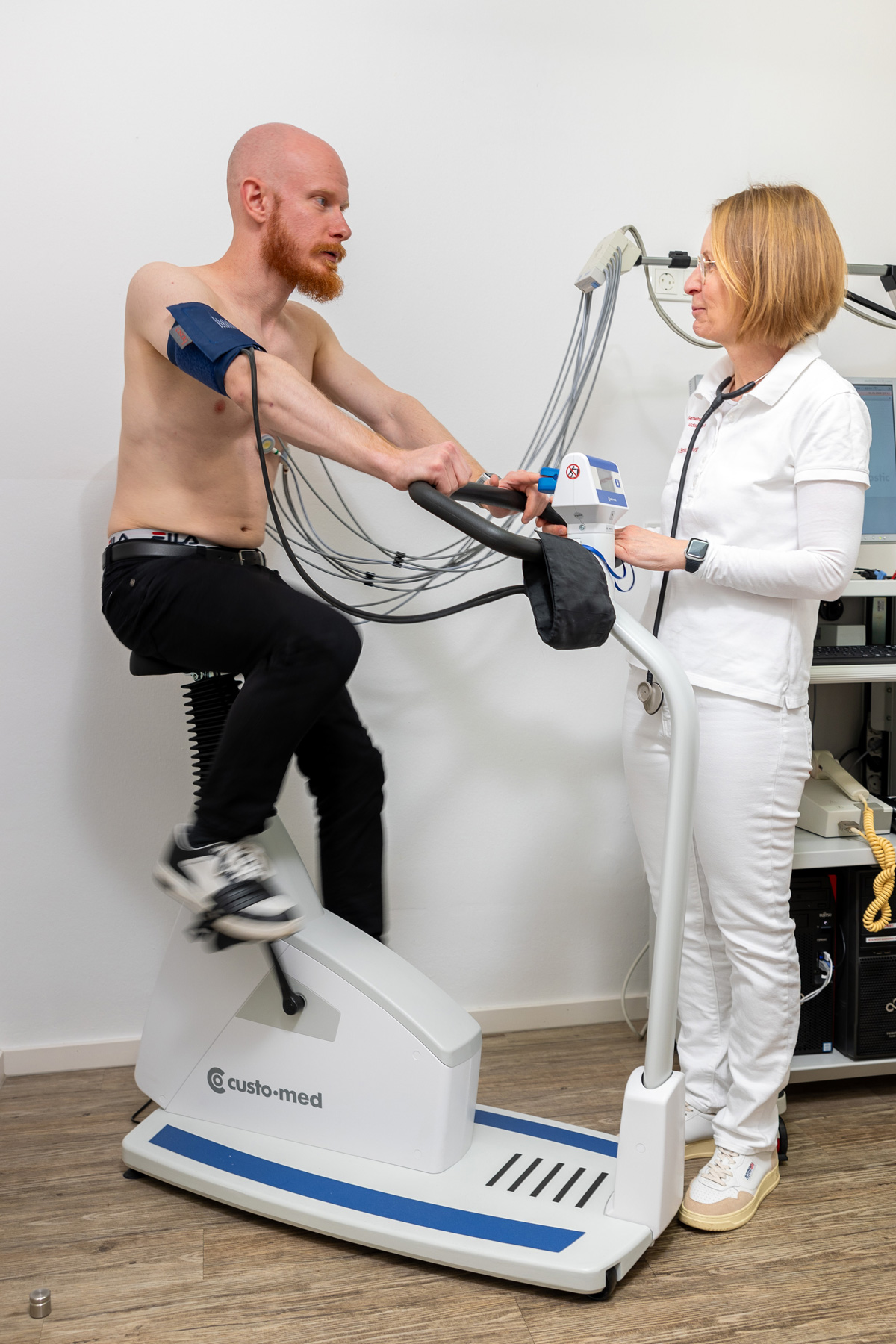 Ein bärtiger Mann trainiert auf einem Ergometer und eine Frau überwacht seine Werte
