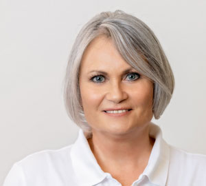 Frau mit kurzen grauen Haaren und hellblauen Augen lächelt freundlich in weißem Hemd