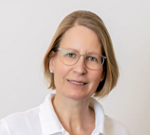 Eine lächelnde Frau mit kurzem blonden Haar und Brille trägt ein weißes Oberteil