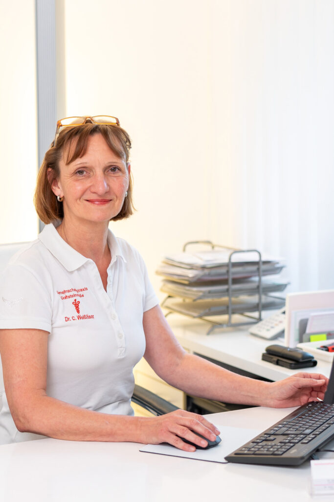 Eine freundliche Frau mittleren Alters mit braunen Haaren sitzt am Schreibtisch in weiss